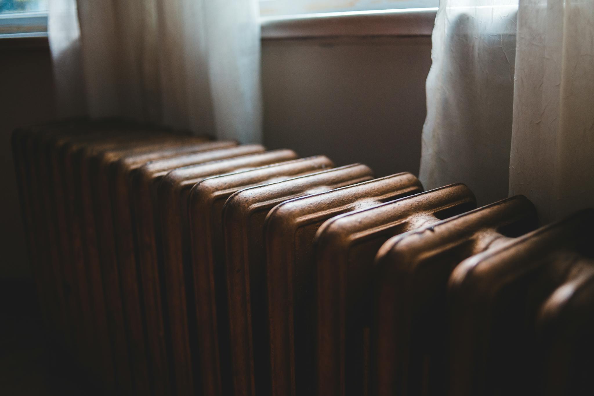 Aged heating battery made of cast iron near windowsill with curtains in house